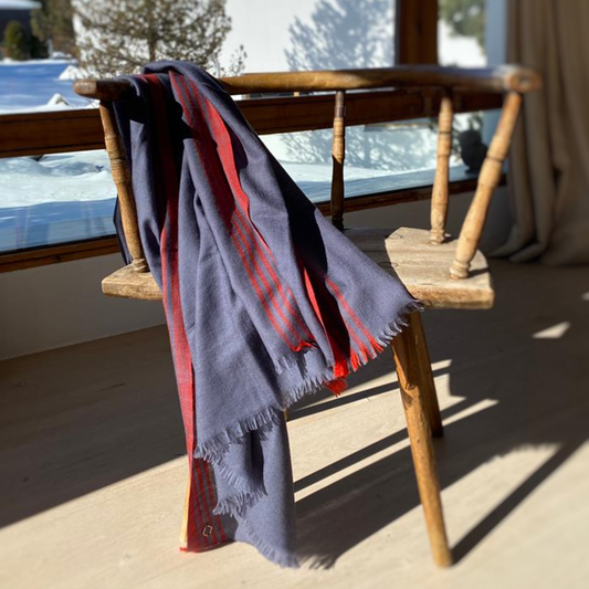 ERUS Kaschmir Schal in Graublau mit roter Streifenborde und kurzen Fransen, über einen Holzstuhl am Fenster drapiert.