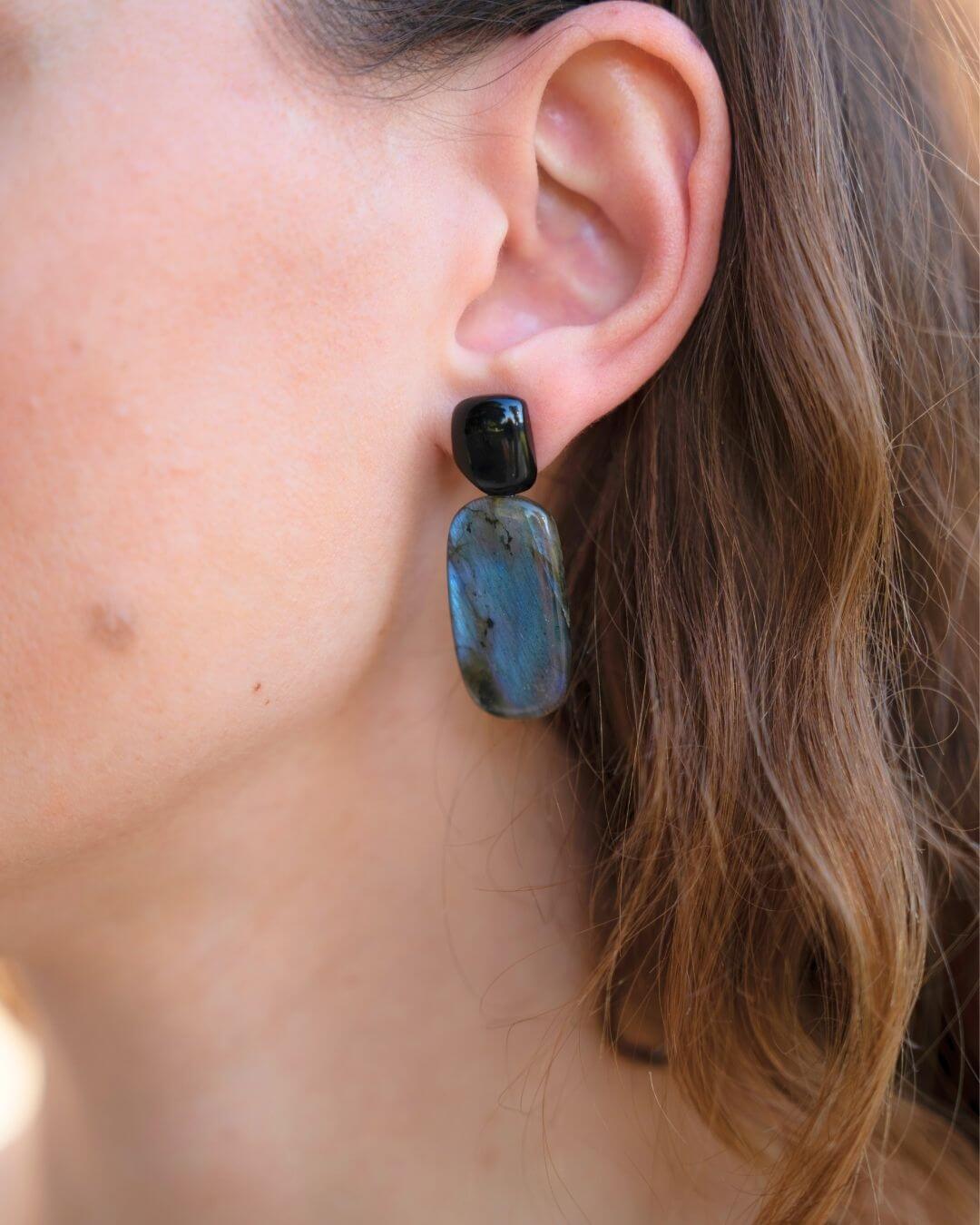 Nahaufnahme eines NYRA Ohrrings mit schwarzem Onyx-Stecker und länglichem Labradorit-Anhänger am Ohr bei offenem Haar.