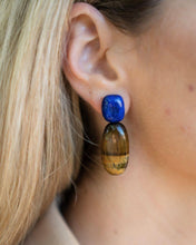 Charger l'image dans la visionneuse de la galerie, PRIYA Ohrring mit blauem Lapislazuli-Stecker und ovalem Tigerauge-Anhänger am Ohr bei offenem blondem Haar.

