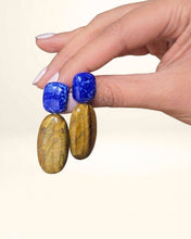 Charger l'image dans la visionneuse de la galerie, PRIYA Ohrringe mit blauem Lapislazuli und goldbraun schimmerndem Tigerauge, in einer Hand gehalten.
