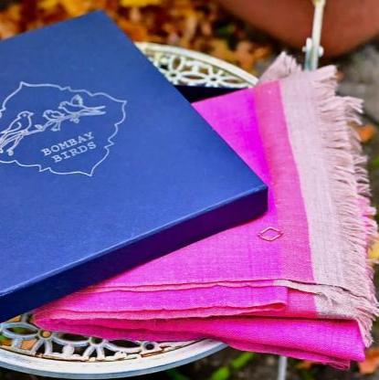 Gefalteter pinkfarbener YADIR Pashmina mit taupefarbenen Fransen neben einer blauen Bombay Birds Geschenkbox.
